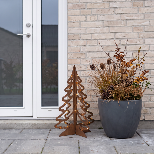 Weihnachtsbaum aus Cortenstahl mit feiner Lichterkette vor einer weißen Haustür neben einem grauen Pflanzkübel.