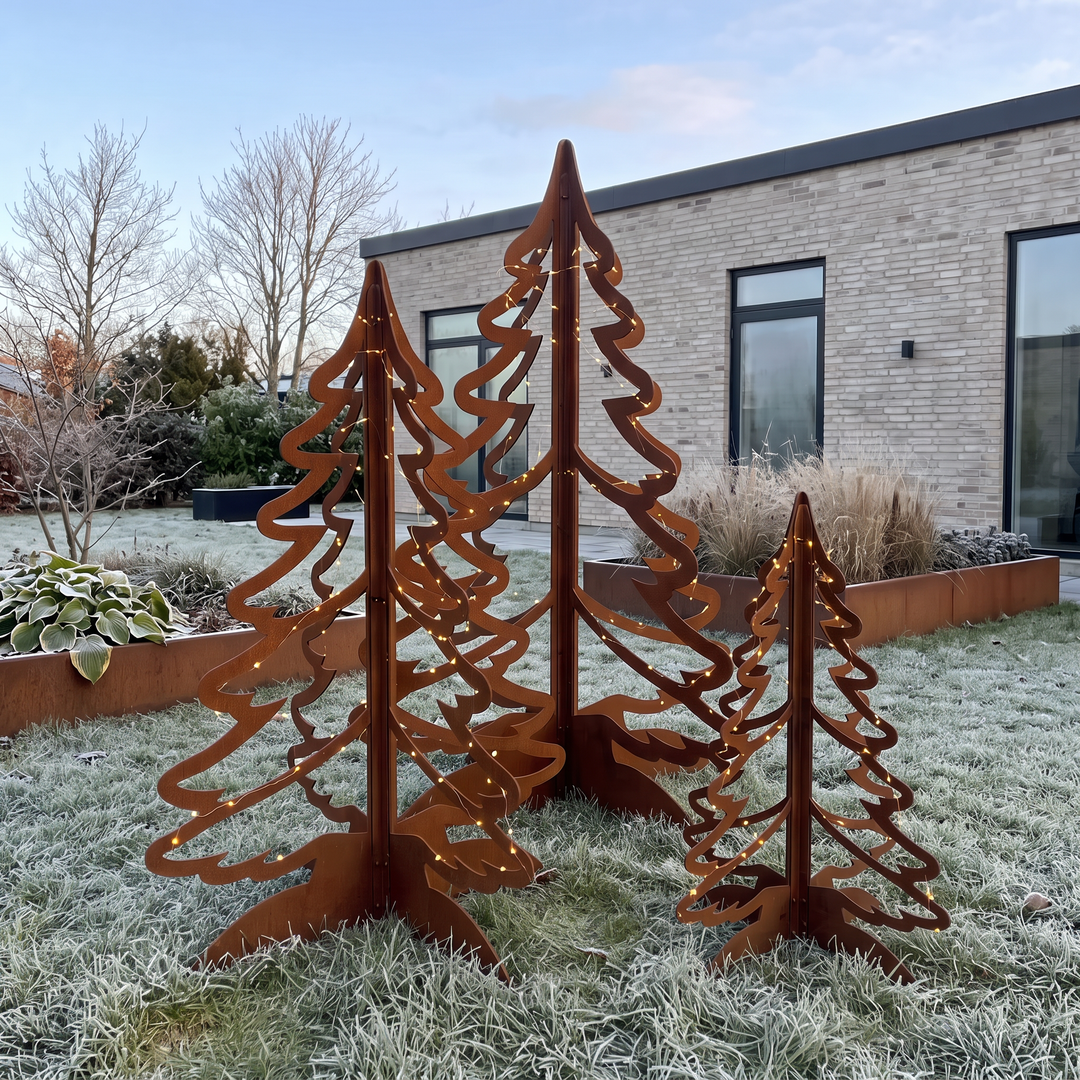 Drei Tannenbäume aus Cortenstahl mit dezenten Lichterketten auf einem frostigen Rasen, dahinter Hochbeete aus Cortenstahl.