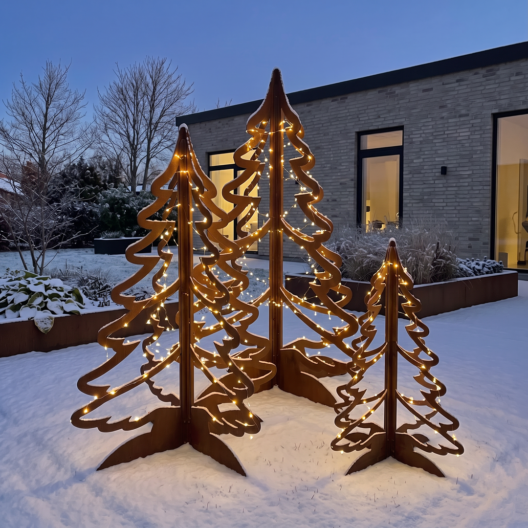 Drei beleuchtete Weihnachtsbäume aus Cortenstahl im verschneiten Garten in der Abenddämmerung vor einem modernen Haus.